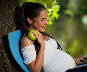 Беременная женщина сидит с ноутбуком и ест яблоко