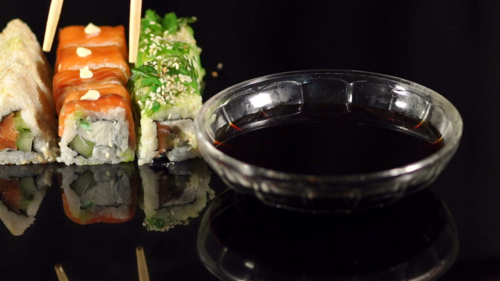 Соевый соус в пиале и суши с палочками на заднем плане на черном фоне
